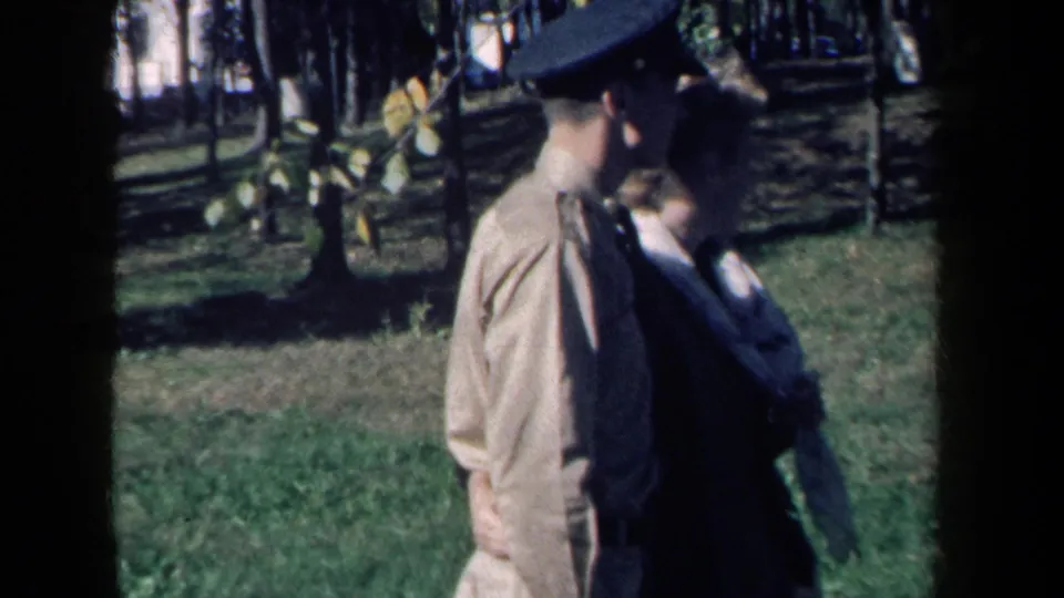 In a New York park scene from 1959, a young uniformed man and a woman walk arm-in-arm across grass under trees.