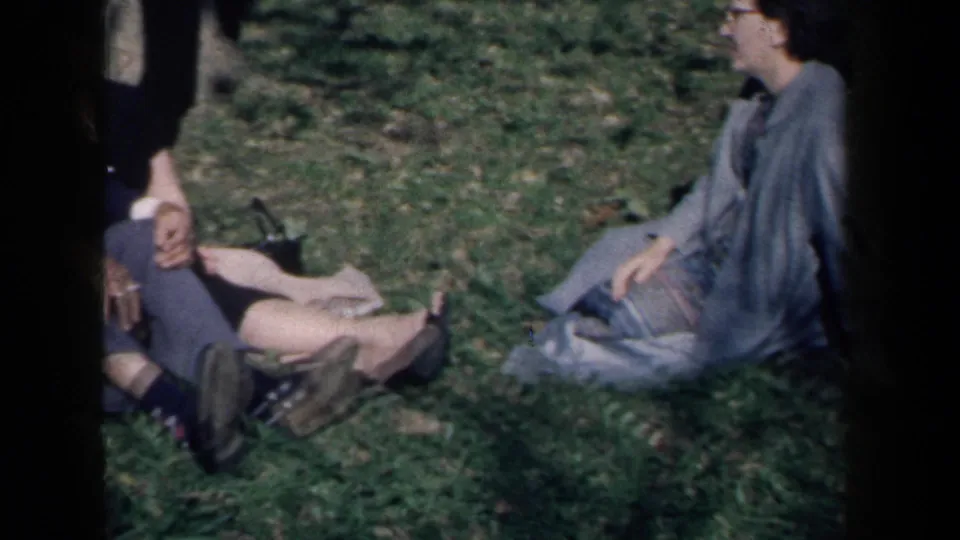 A cropped 1959 New York home-movie frame shows adults seated on grass, with the camera partly blocked by contact-sheet borders.