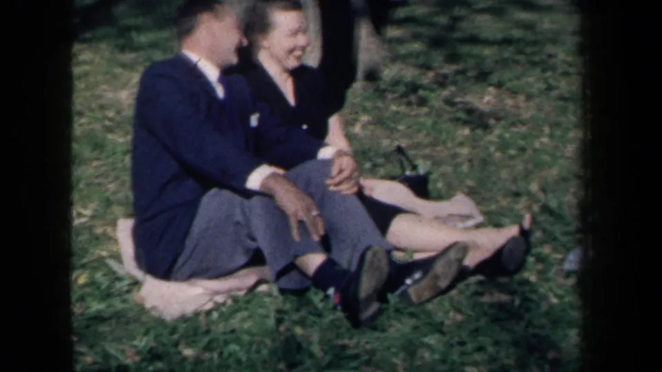 A smiling couple sits close together on a grassy lawn in the supported 1959 New York home-movie scene.