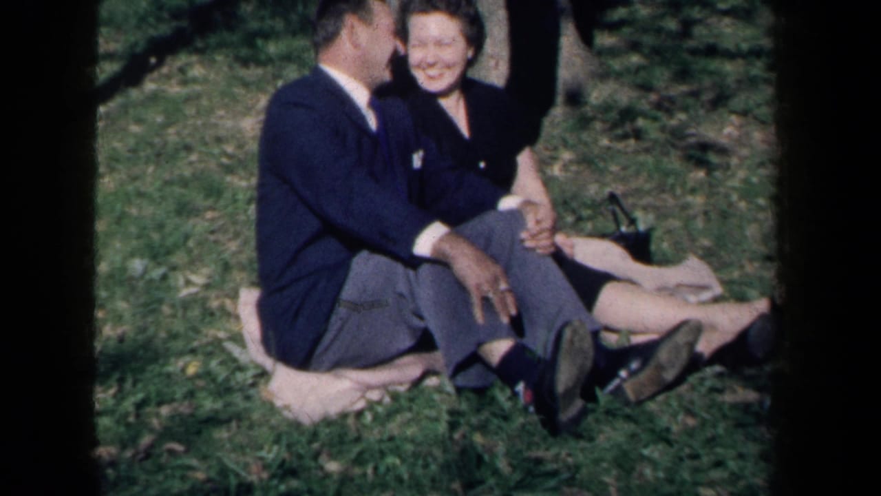 In 1959 New York home-movie footage, a smiling adult couple sits close together on a light blanket in grass.