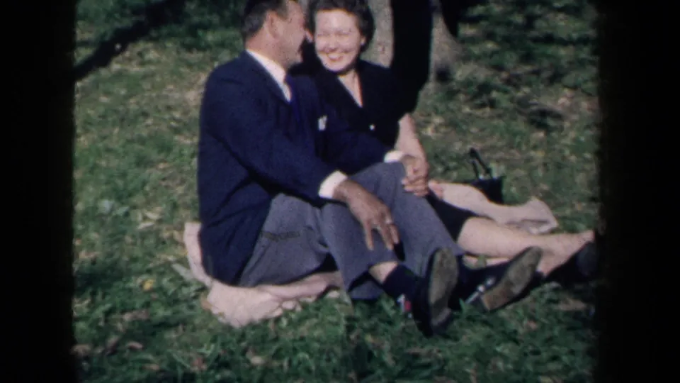 In 1959 New York home-movie footage, a smiling adult couple sits close together on a light blanket in grass.