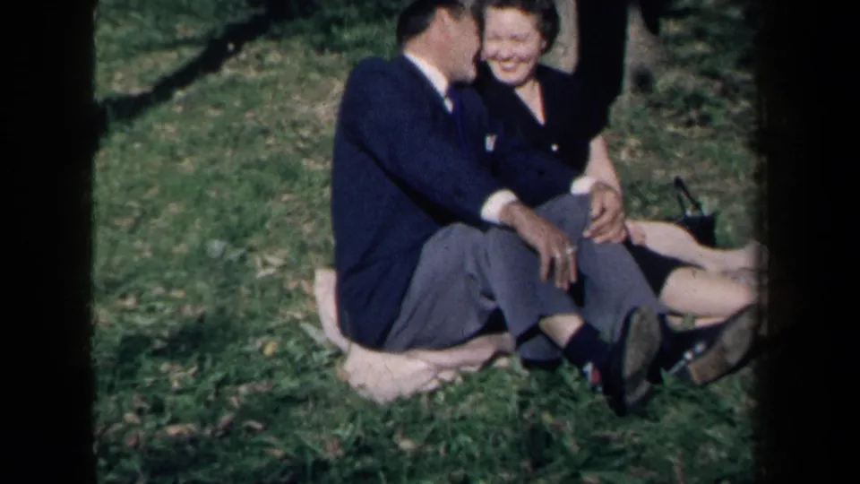 The couple remains seated on a blanket in the park, leaning close as the woman smiles toward the camera.