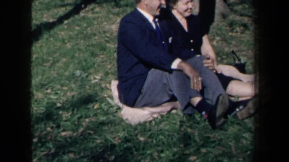 The adult couple sits side by side on a light blanket in a grassy New York park, with a small dark handbag nearby.
