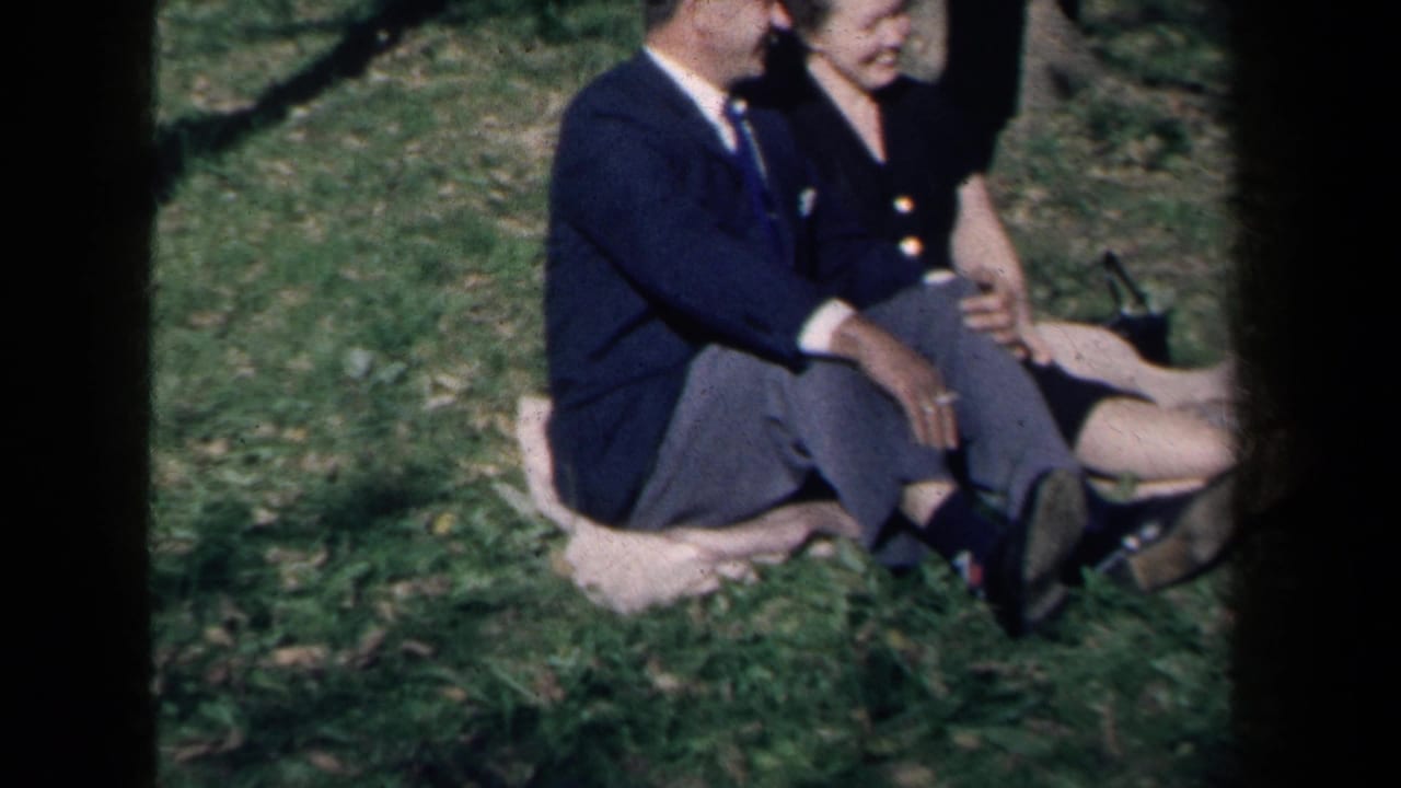 Adults seated on a grassy lawn, with a suited man and a woman in a dark dress visible through 8mm frame edges.
