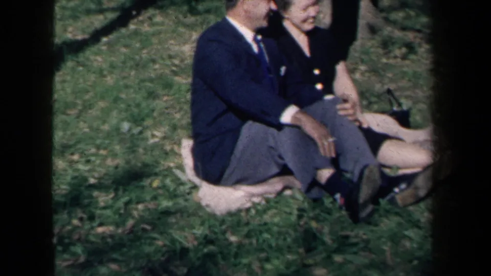 1959 New York home-movie frame showing adults seated on grass, including a suited man and a woman in a dark dress, partly obscured by film borders.