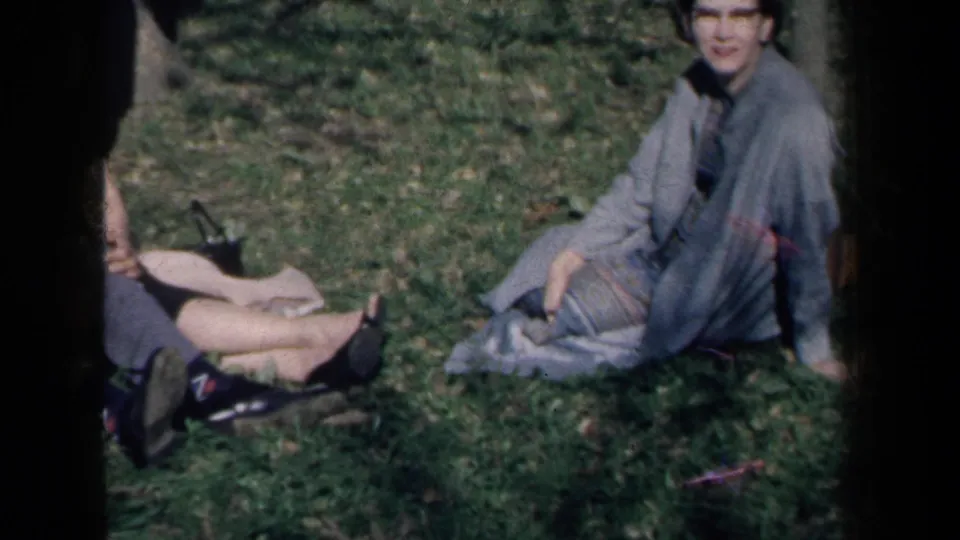 1959 New York home-movie frame showing a woman in a light gray coat seated on grass near another adult's legs and shoes.