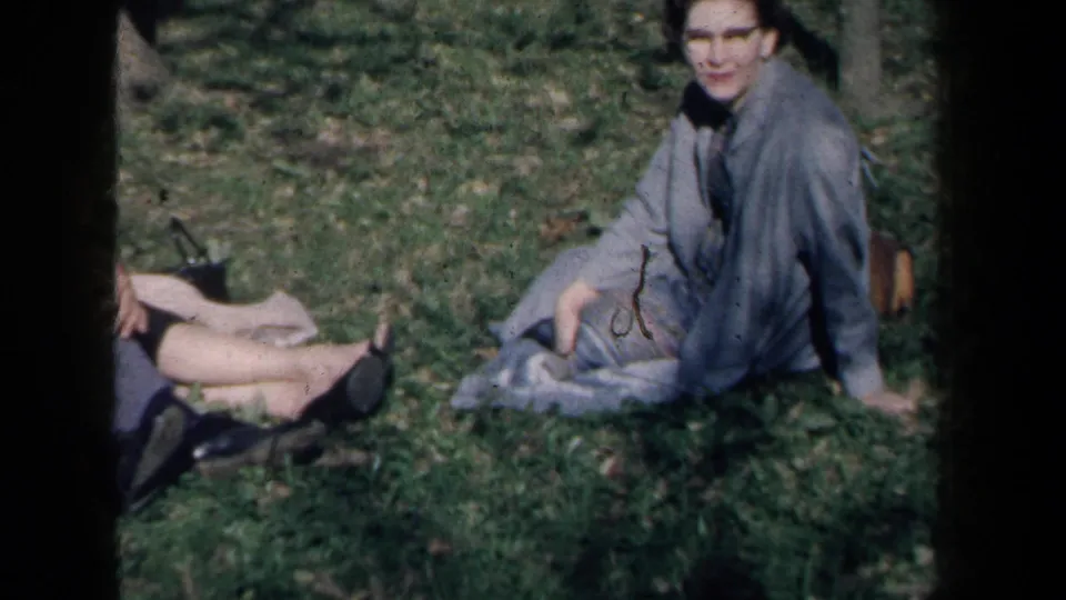 1959 New York home-movie frame of a woman in a light gray coat seated on grass, with another adult partly visible nearby.