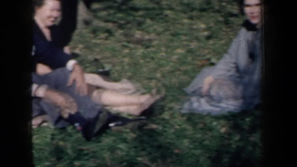 1959 New York 8mm frame showing adults seated on grass, including a couple on a blanket and a woman nearby in a light dress.