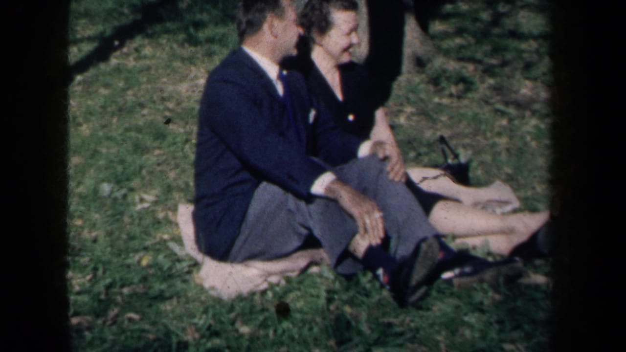 1959 New York home-movie frame of a suited man and smiling woman seated together on a blanket in a grassy park.