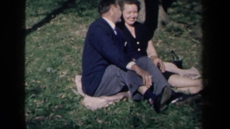 1959 New York 8mm frame with a smiling woman in a dark dress and a suited man seated closely on a blanket.