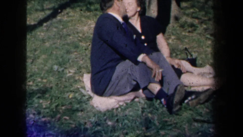 1959 New York home-movie frame of a suited man leaning toward a woman on a park blanket, with grass and trees behind them.