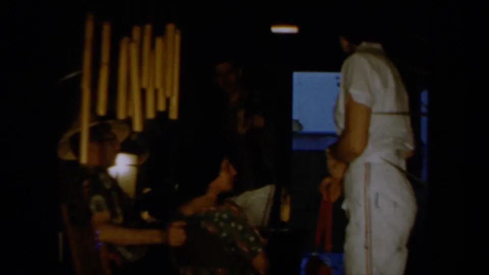1961 California home-movie frame showing seated party guests in tropical hats with a person in a white outfit standing in a dark interior.