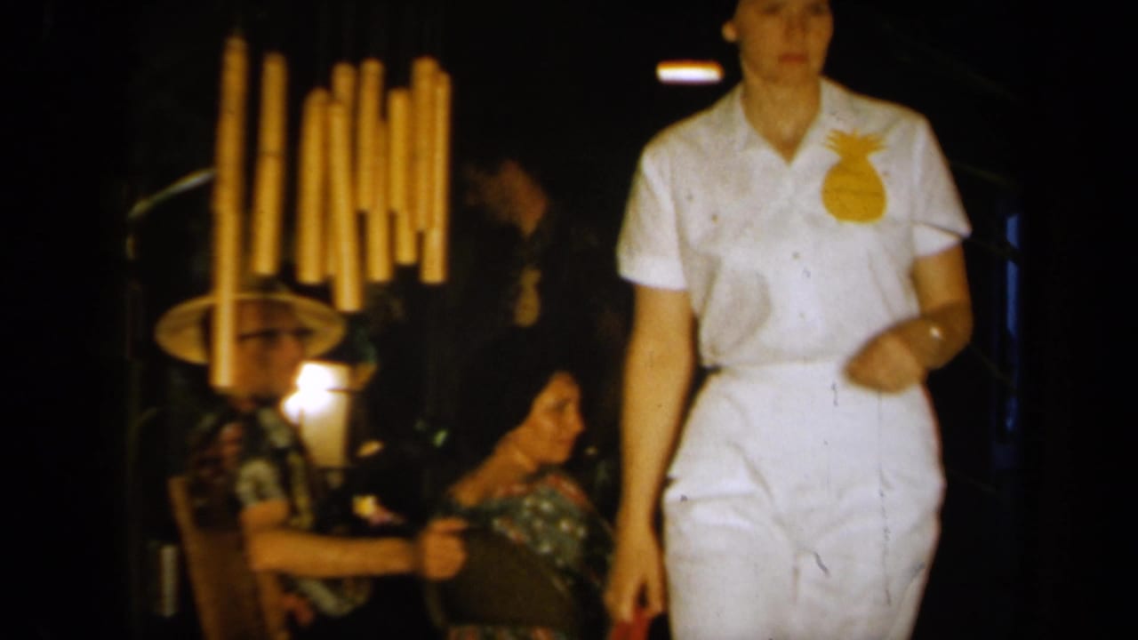 1961 California party frame with a white-clad figure wearing a pineapple emblem passing seated guests in tropical hats.