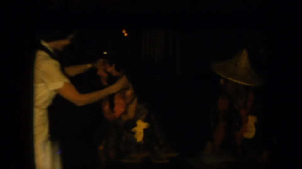 1961 California home-movie frame showing a partygoer placing a flower lei on a seated guest during a Polynesian-themed gathering.