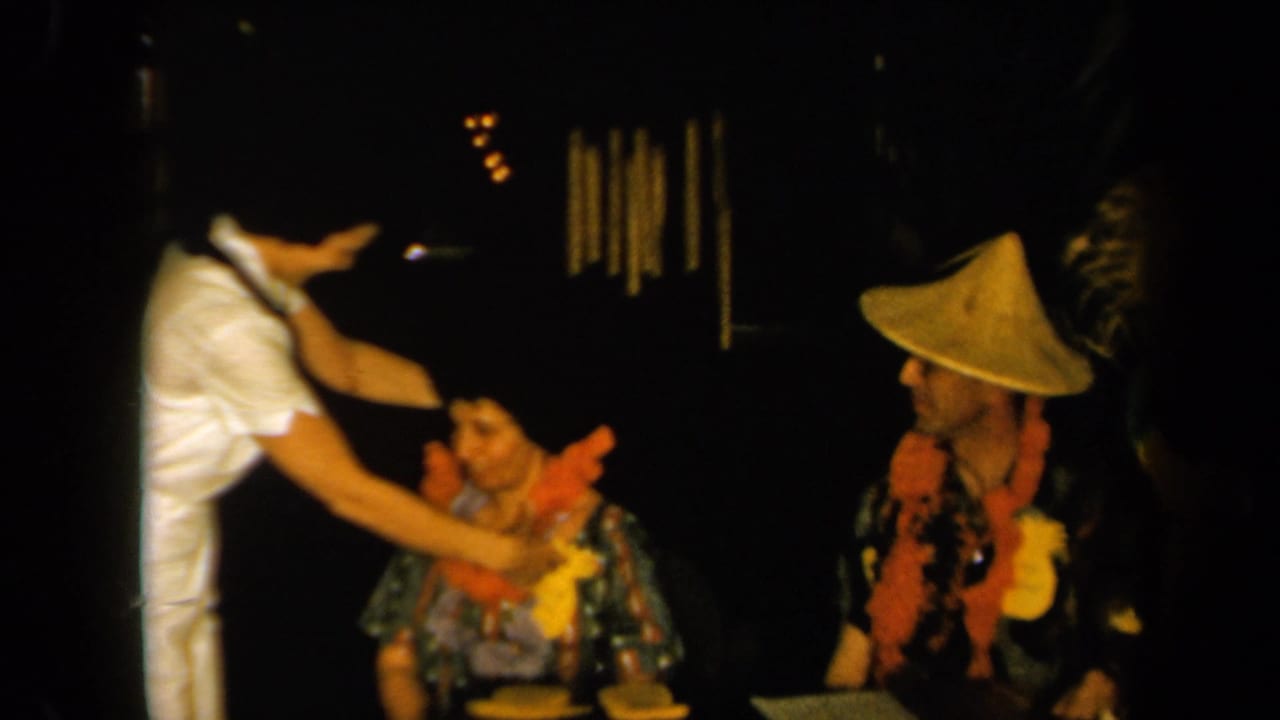 1961 California 8mm frame showing a woman in white adjusting or placing a lei on a seated woman as a man in a conical hat observes nearby.