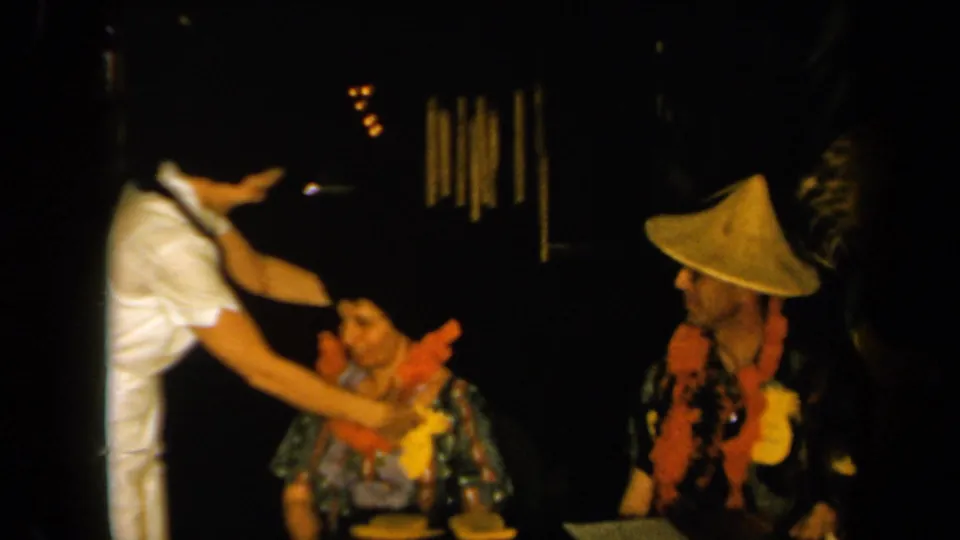 1961 California 8mm frame showing a woman in white adjusting or placing a lei on a seated woman as a man in a conical hat observes nearby.