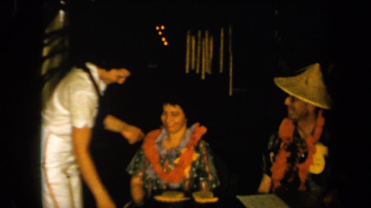 1961 California home-movie frame with a seated woman in a patterned dress and orange lei beside a man in a conical hat and lei.