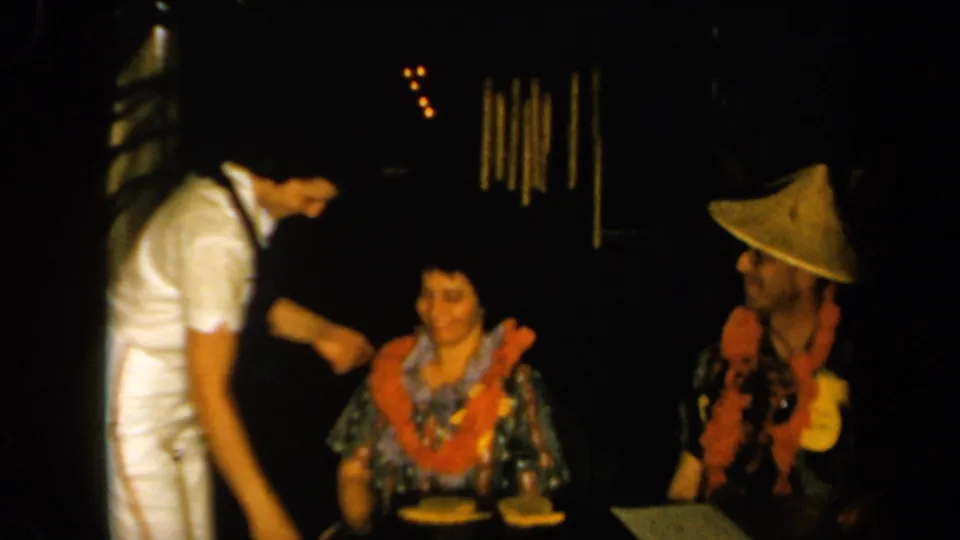 1961 California home-movie frame with a seated woman in a patterned dress and orange lei beside a man in a conical hat and lei.