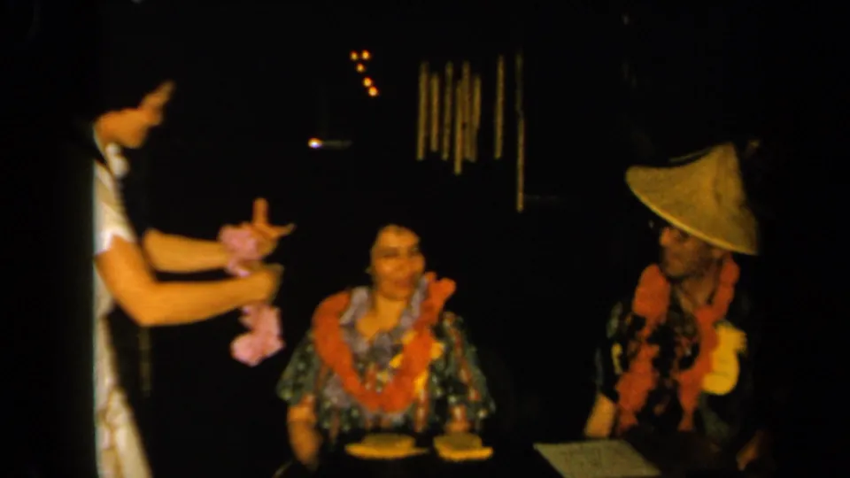 1961 California 8mm frame showing a woman in white presenting a pink floral lei near a seated woman at a dimly lit table.