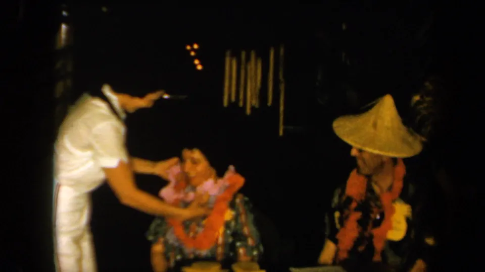 1961 California home-movie frame showing a pink lei being placed on a seated woman during a dim nighttime social gathering.