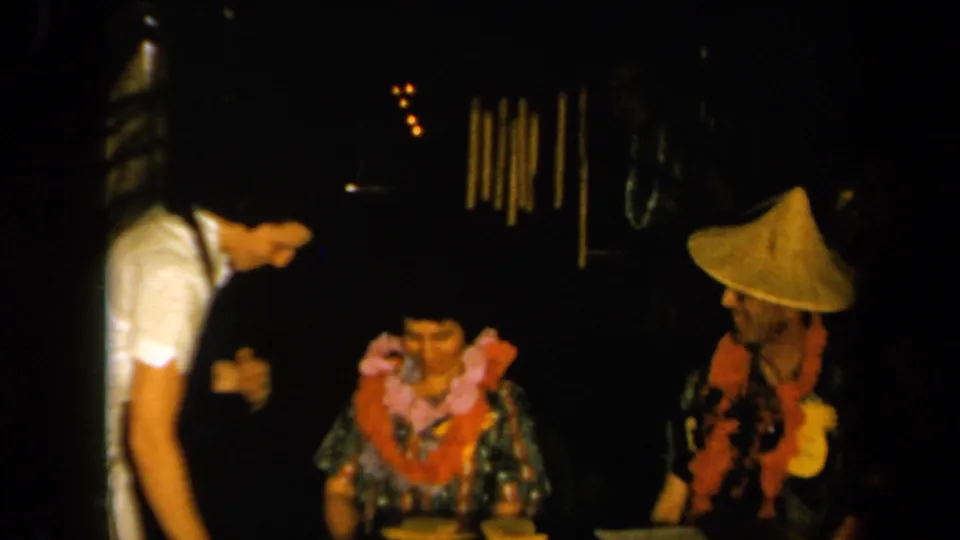 1961 California home-movie frame of seated dinner guests in leis, with a standing person leaning toward the table in a dim interior.