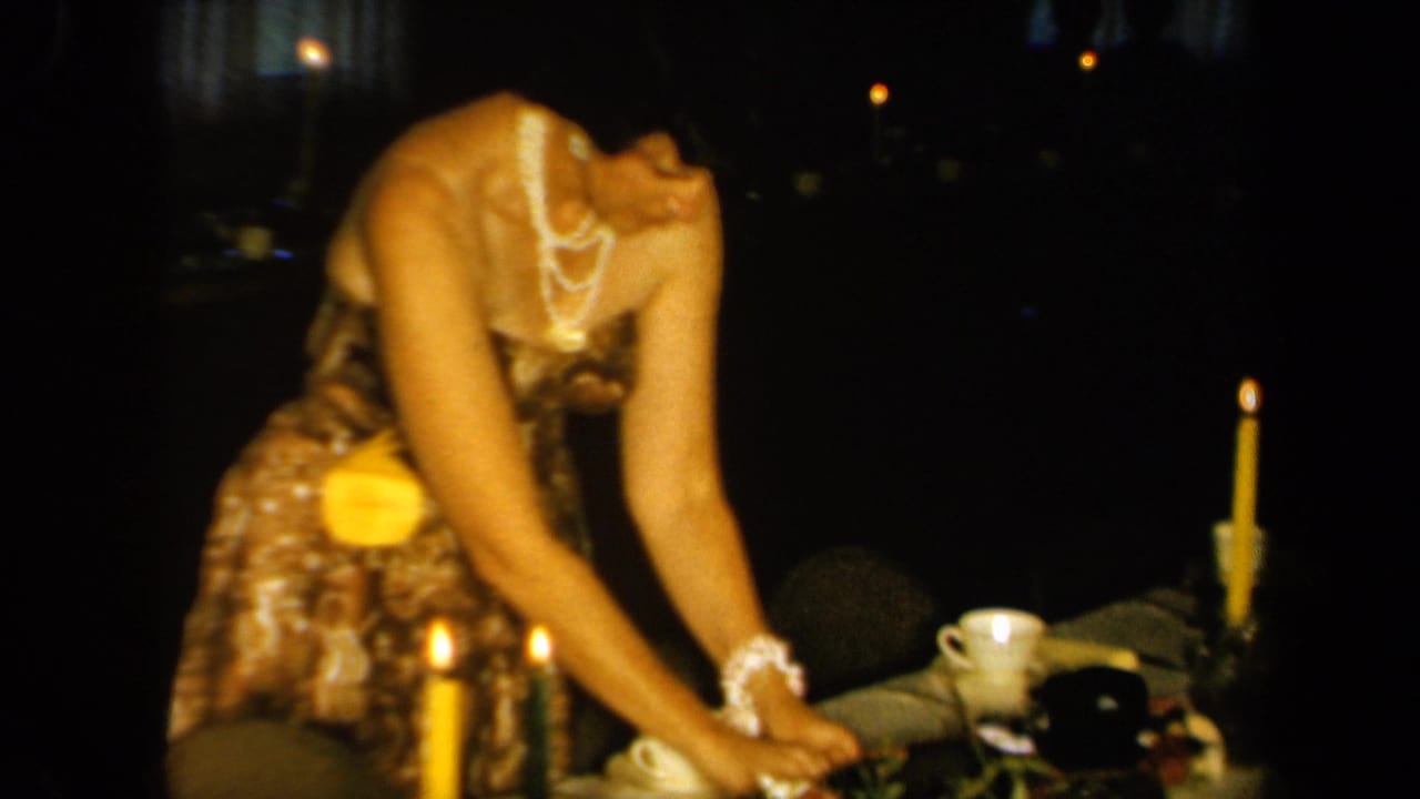 1961 California home-movie frame showing a woman in a patterned dress and pearls arranging items on a candlelit table.