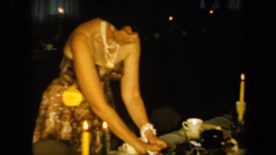 1961 California home-movie frame showing a woman in a patterned dress and pearls arranging items on a candlelit table.