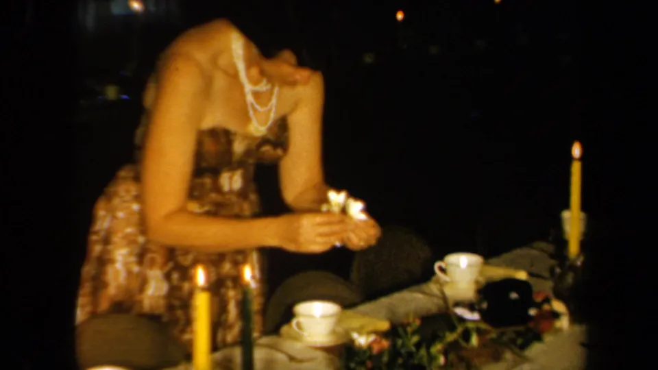 In 1961 California home-movie footage, a woman in a patterned dress and pearl necklace arranges flowers on a candlelit table.