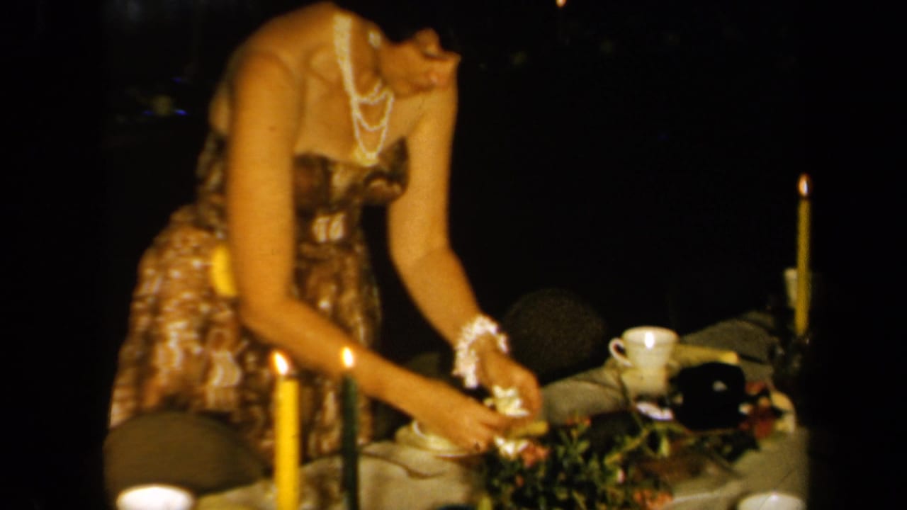 The woman reaches over a candlelit Hawaiian-themed table arrangement, with cups and floral decorations visible in the 1961 California scene.