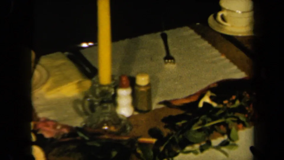 1961 California home-movie view of a candlelit table with glass holder, small shakers, fork, folded napkin, and flowers.