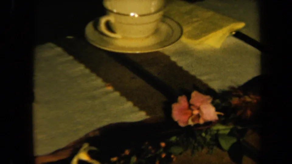 1961 California home-movie frame showing a cup and saucer on a decorated table with folded napkin and pink flowers.