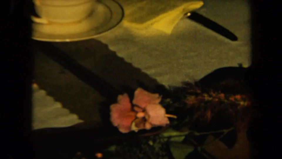 Warm 1961 8mm California home-movie frame showing a teacup, saucer, knife, napkin, and pink flower on a dark table.