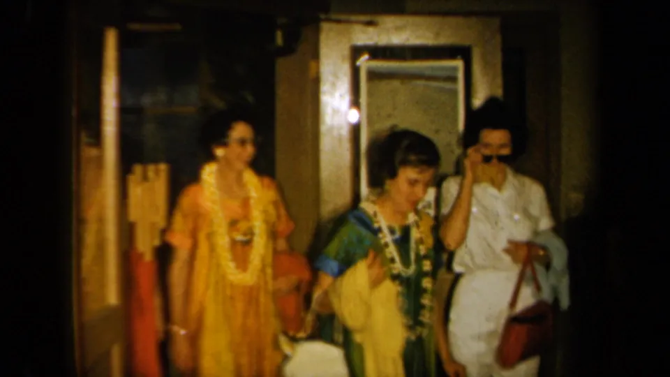 Women at a 1961 California indoor gathering wear leis and colorful clothing near a doorway, mirror, and framed wall art.