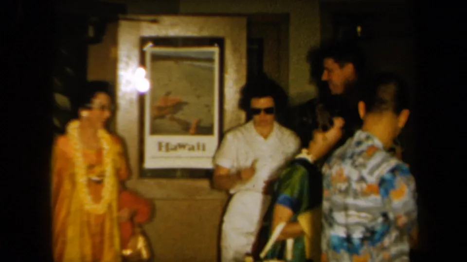 A 1961 California home-movie frame shows party guests in leis near a framed Hawaii poster and warm interior lighting.