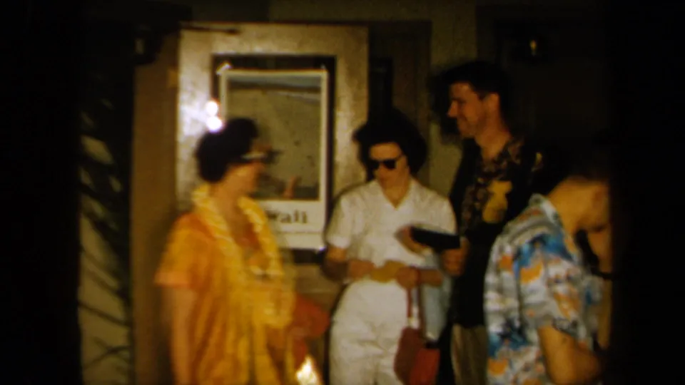 1961 California home-movie frame showing adults at a Hawaiian-themed indoor party, with leis, sunglasses, and a Hawaii poster visible.