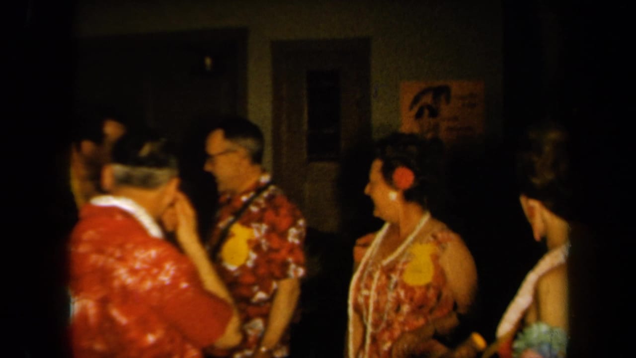 1961 California home-movie frame of adults conversing at a Hawaiian-themed party, with leis, aloha shirts, and private interior details.
