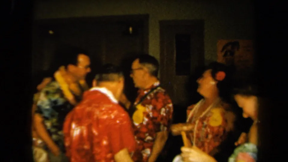 1961 California home-movie frame showing adults in leis and bright patterned clothing socializing at a Hawaiian-themed indoor gathering.
