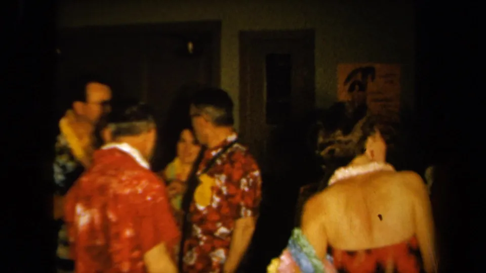 1961 California home-movie frame of a luau-themed indoor party, with adults in leis, patterned shirts, and a woman seen from behind.