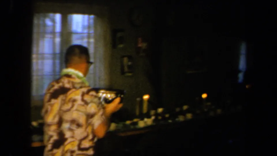 A man in a patterned shirt carries a camera near a table with candles during the 1961 California home party.