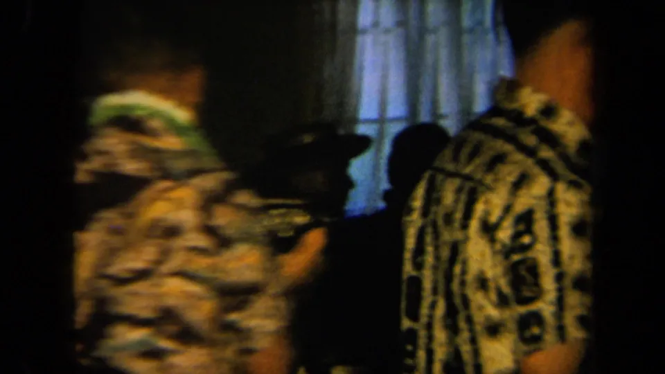 Two men in Hawaiian-style shirts move through a dim 1961 California party room, one partly blurred by handheld motion.