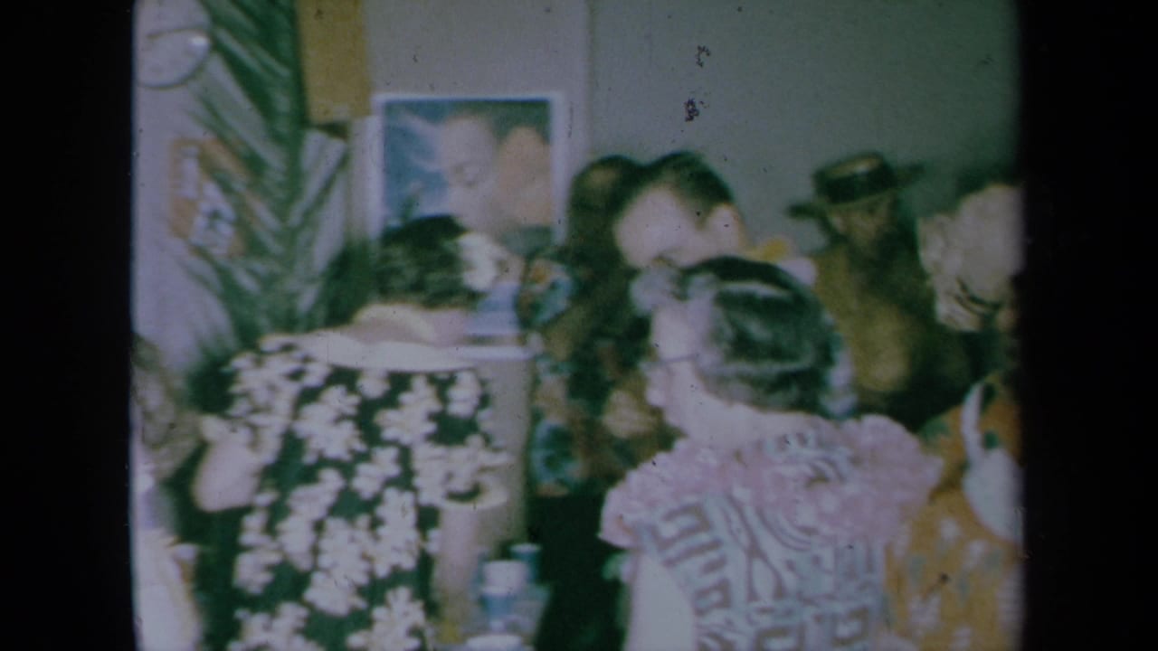 Adults gather closely in a private room, with floral dresses, glasses, wall art, and a plant visible.