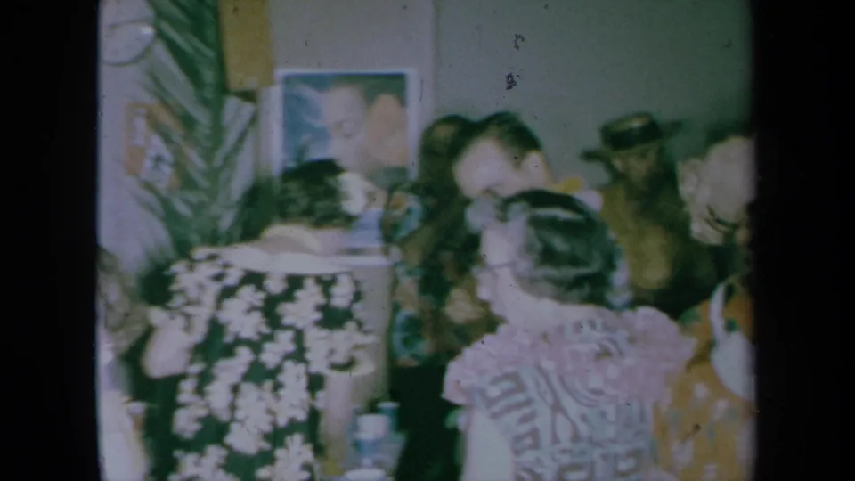 1961 California home-movie frame showing adults mingling indoors at a private house party, with floral clothing, cups, framed art, and a plant.