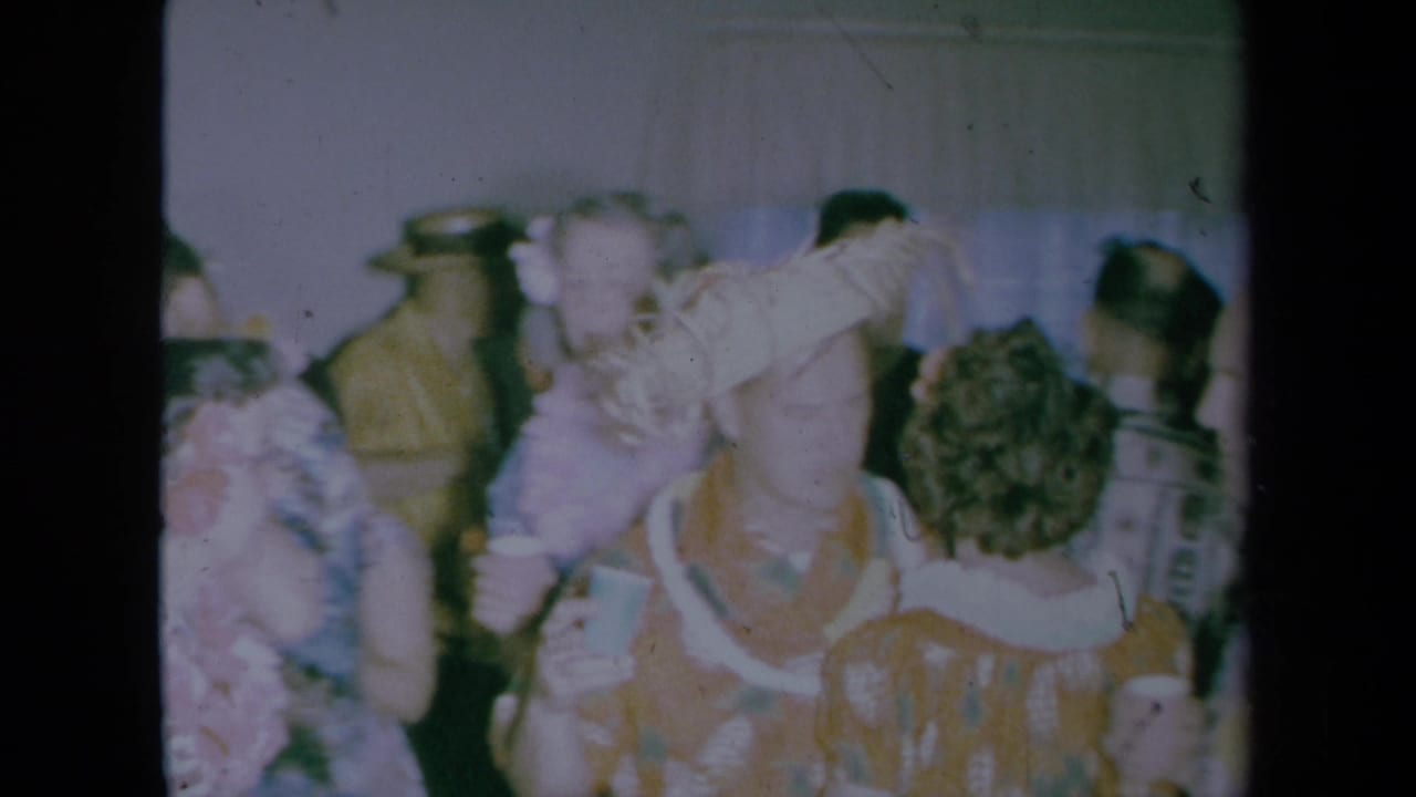 Adults in party clothing and hats mingle indoors, with cups and wall details visible behind them.