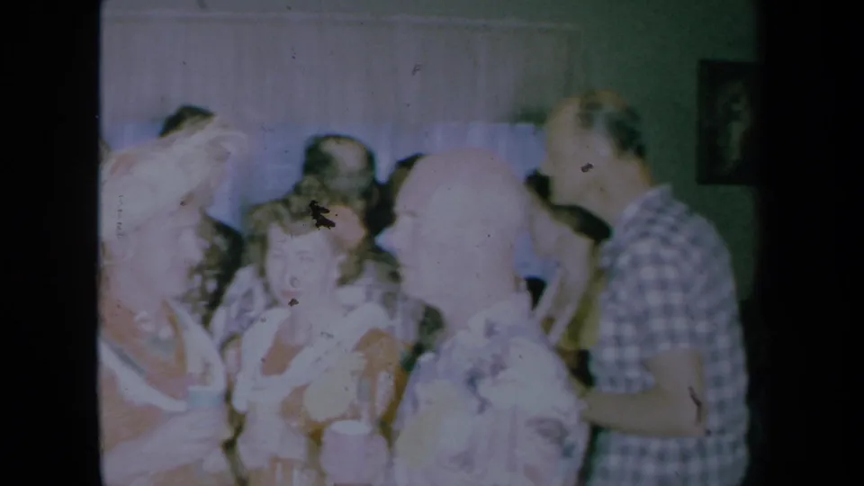 1961 California home-movie frame of adults talking at a crowded house party, with plaid shirt, light dresses, cups, and framed wall art visible.
