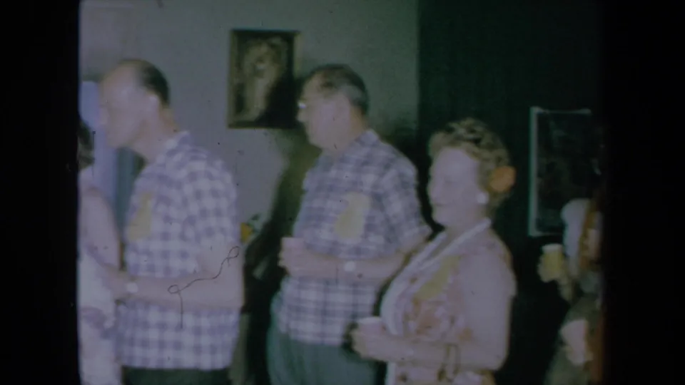 Adults at a 1961 California house party stand with drinks near framed wall pictures, captured in soft, faded 8mm color.