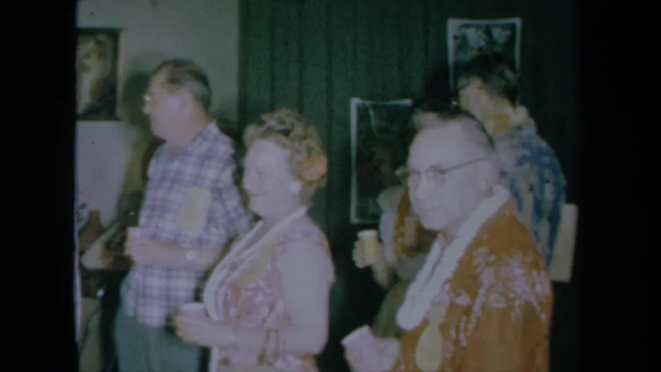 California 1961 home-movie frame showing adult party guests in patterned and plaid clothing standing indoors with drinks.
