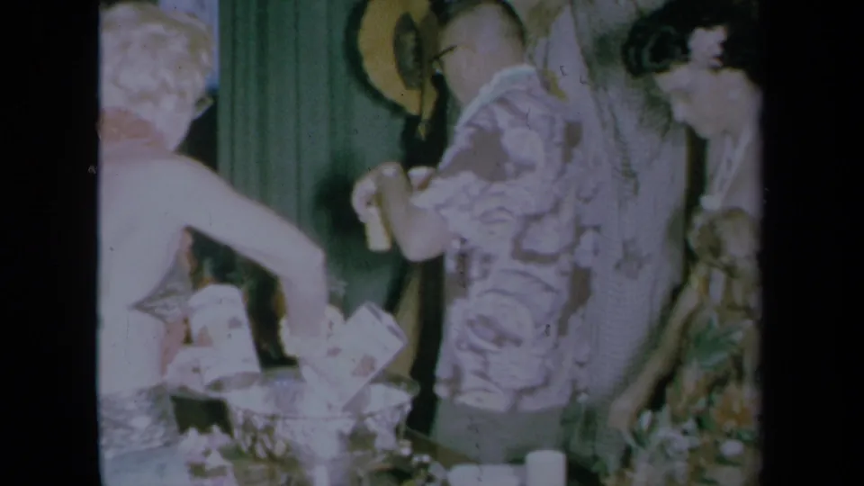 1961 California home-movie frame showing adults serving drinks by a punch bowl in a private indoor party setting.