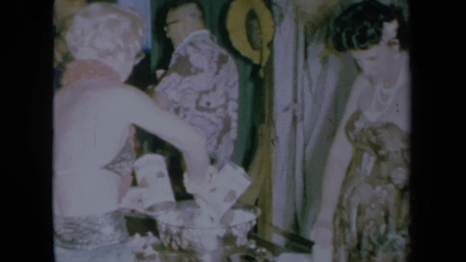 California 1961 indoor party frame with a woman in a patterned dress near the punch bowl and other adults in the background.
