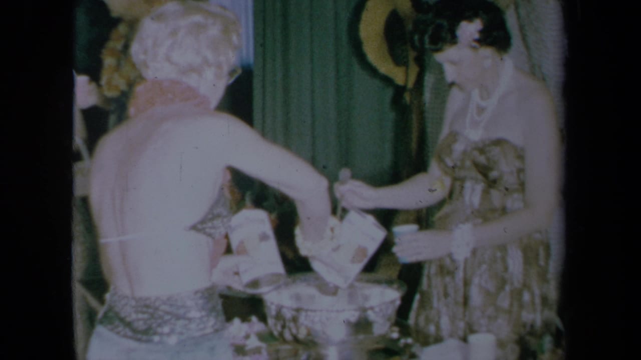 Two women in party dresses work around a punch bowl in a 1961 California home-movie interior.