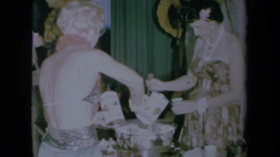 Two women in party dresses work around a punch bowl in a 1961 California home-movie interior.